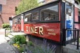 A classic diner with a red and black exterior and large windows is situated next to a well-maintained garden with various colorful flowers. The word 'DINER' is prominently displayed in bold yellow letters, alongside the phrase 'LADIES INVITED.' There is a door with an 'OPEN' sign and a glimpse of the interior can be seen through the windows.