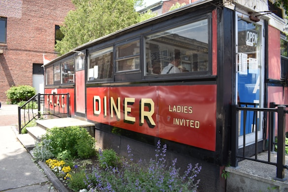 A classic diner with a red and black exterior and large windows is situated next to a well-maintained garden with various colorful flowers. The word 'DINER' is prominently displayed in bold yellow letters, alongside the phrase 'LADIES INVITED.' There is a door with an 'OPEN' sign and a glimpse of the interior can be seen through the windows.