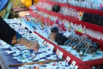 A tabletop display at a market features a variety of colorful glass figurines, ceramic items, and small model motorcycles. The items are spread across tiered stands with a fabric cover, and a person's hand is reaching towards the table, suggesting browsing. Numerous small cards are positioned in front of the items.