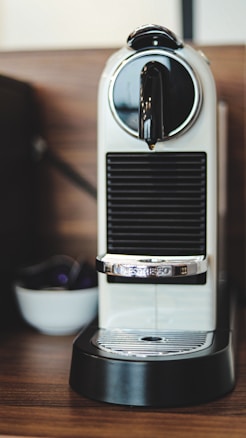 A sleek, modern espresso machine with a rounded top and chrome accents is placed on a wooden countertop. The machine has a white and black color scheme with a prominent chrome handle. In the background, there is a small bowl that appears to hold coffee pods.