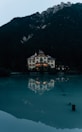 A large, elegant house with traditional architecture is situated by the edge of a tranquil body of water, reflecting its illuminated windows in the surface. Surrounded by dense forest, the house is set against a backdrop of towering mountains. The overall scene is calm and picturesque, with a quiet, serene atmosphere.