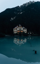 A large, elegant house with traditional architecture is situated by the edge of a tranquil body of water, reflecting its illuminated windows in the surface. Surrounded by dense forest, the house is set against a backdrop of towering mountains. The overall scene is calm and picturesque, with a quiet, serene atmosphere.