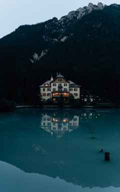 A large, elegant house with traditional architecture is situated by the edge of a tranquil body of water, reflecting its illuminated windows in the surface. Surrounded by dense forest, the house is set against a backdrop of towering mountains. The overall scene is calm and picturesque, with a quiet, serene atmosphere.