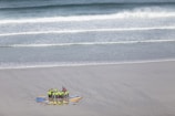 A group of people wearing bright green and red clothing gather in a circle on a sandy beach, surrounded by colorful surfboards arranged like a sunburst. The ocean waves gently roll towards the shore in the background.