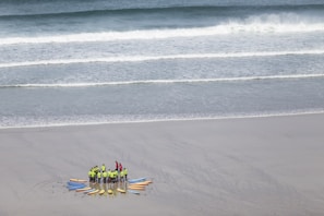 A group of people wearing bright green and red clothing gather in a circle on a sandy beach, surrounded by colorful surfboards arranged like a sunburst. The ocean waves gently roll towards the shore in the background.