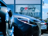 Several cars are parked in front of a dealership with a large window displaying the words 'Buy happy' and 'Brown's Fairfax Mazda'. The focus is on the cars in the foreground, while the dealership building is partially visible in the background.