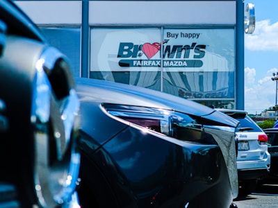 Several cars are parked in front of a dealership with a large window displaying the words 'Buy happy' and 'Brown's Fairfax Mazda'. The focus is on the cars in the foreground, while the dealership building is partially visible in the background.