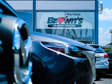 Several cars are parked in front of a dealership with a large window displaying the words 'Buy happy' and 'Brown's Fairfax Mazda'. The focus is on the cars in the foreground, while the dealership building is partially visible in the background.
