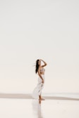 A woman is standing on a serene beach, wearing a bikini top and a flowing white skirt. Her hand is raised to her forehead as she gazes into the distance. The setting has a peaceful and reflective ambiance, with a soft, muted color palette.