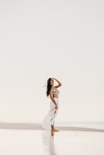 A woman is standing on a serene beach, wearing a bikini top and a flowing white skirt. Her hand is raised to her forehead as she gazes into the distance. The setting has a peaceful and reflective ambiance, with a soft, muted color palette.