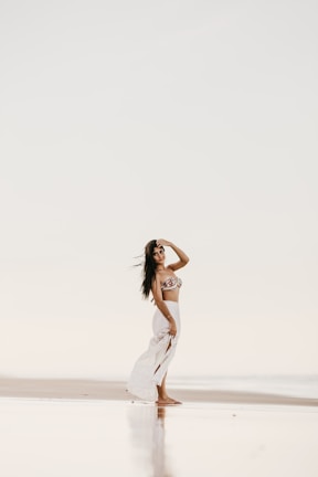A woman is standing on a serene beach, wearing a bikini top and a flowing white skirt. Her hand is raised to her forehead as she gazes into the distance. The setting has a peaceful and reflective ambiance, with a soft, muted color palette.