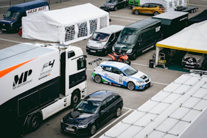 A motorsport paddock filled with support vehicles and race cars. A large white and black truck with 'MP Motorsport' branding is adjacent to a white and blue race car numbered 36. Multiple vans and support tents are visible, indicating a professional racing setup.