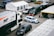 A motorsport paddock filled with support vehicles and race cars. A large white and black truck with 'MP Motorsport' branding is adjacent to a white and blue race car numbered 36. Multiple vans and support tents are visible, indicating a professional racing setup.