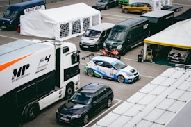 A motorsport paddock filled with support vehicles and race cars. A large white and black truck with 'MP Motorsport' branding is adjacent to a white and blue race car numbered 36. Multiple vans and support tents are visible, indicating a professional racing setup.