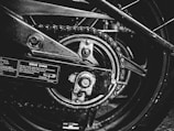 A close-up black and white photograph of a motorcycle's drive chain and sprocket. The image captures the intricacies of the mechanical components, including the chain links, sprocket teeth, and metal bolts. Labels with instructions and measurements are visible on the metallic surface, emphasizing the industrial nature of the scene.