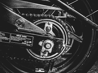 A close-up black and white photograph of a motorcycle's drive chain and sprocket. The image captures the intricacies of the mechanical components, including the chain links, sprocket teeth, and metal bolts. Labels with instructions and measurements are visible on the metallic surface, emphasizing the industrial nature of the scene.