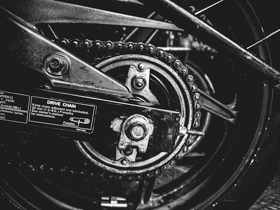 A close-up black and white photograph of a motorcycle's drive chain and sprocket. The image captures the intricacies of the mechanical components, including the chain links, sprocket teeth, and metal bolts. Labels with instructions and measurements are visible on the metallic surface, emphasizing the industrial nature of the scene.