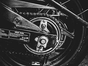 A close-up black and white photograph of a motorcycle's drive chain and sprocket. The image captures the intricacies of the mechanical components, including the chain links, sprocket teeth, and metal bolts. Labels with instructions and measurements are visible on the metallic surface, emphasizing the industrial nature of the scene.