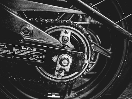 A close-up black and white photograph of a motorcycle's drive chain and sprocket. The image captures the intricacies of the mechanical components, including the chain links, sprocket teeth, and metal bolts. Labels with instructions and measurements are visible on the metallic surface, emphasizing the industrial nature of the scene.