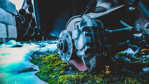 A close-up of a rusty car brake rotor and caliper, surrounded by green moss and patches of snow. In the background, there are stacked metal pipes and concrete blocks, suggesting an industrial setting.