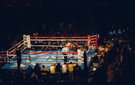 A boxing match is taking place in a crowded arena. The ring is surrounded by photographers and judges, and the audience fills the background. Bright overhead lights illuminate the boxing ring, highlighting the fighters. Ringside banners and advertisements decorate the area.