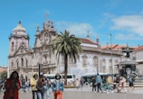 A historic church with ornate architecture and intricate details is surrounded by people walking and engaging with their phones. A large palm tree sits in front of the church, and buses are visible in the background. A decorative fountain is also present, adding to the charm of the square.