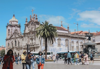 A historic church with ornate architecture and intricate details is surrounded by people walking and engaging with their phones. A large palm tree sits in front of the church, and buses are visible in the background. A decorative fountain is also present, adding to the charm of the square.