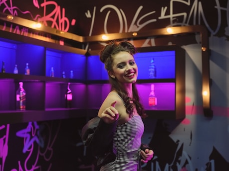 A woman with brown hair styled in an updo, wearing a checkered dress, smiles and points toward the camera. She stands in a lively, colorful setting with neon lighting in shades of pink and blue. The background features shelves with bottles and graffiti-style decorations on the walls, creating an eclectic and vibrant atmosphere.