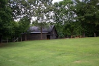 A rustic wooden barn nestled among tall, lush trees, surrounded by a well-kept green lawn. The structure has a weathered appearance, indicating age and use, and is partially shaded by the overhanging branches of the trees.