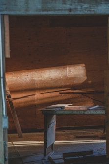 Warm light casts across a rustic wooden interior, illuminating a simple desk with papers scattered on top. The wooden panels and muted tones create a serene and contemplative atmosphere.