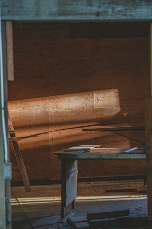 Warm light casts across a rustic wooden interior, illuminating a simple desk with papers scattered on top. The wooden panels and muted tones create a serene and contemplative atmosphere.