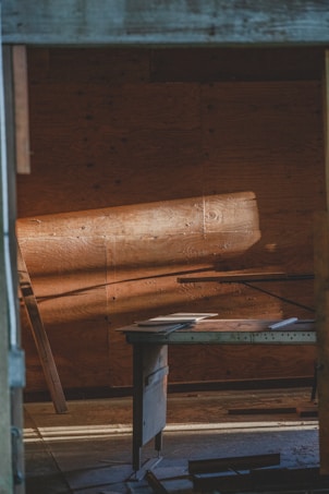 Warm light casts across a rustic wooden interior, illuminating a simple desk with papers scattered on top. The wooden panels and muted tones create a serene and contemplative atmosphere.