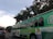 A large green trolley-style bus with multiple windows is seen on a city street. It bears signage promoting walking and biking safety, with a colorful design including images of people walking. A silver car is visible in the lane next to the bus, and there are traffic lights and trees in the background under a partly cloudy sky.