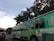 A large green trolley-style bus with multiple windows is seen on a city street. It bears signage promoting walking and biking safety, with a colorful design including images of people walking. A silver car is visible in the lane next to the bus, and there are traffic lights and trees in the background under a partly cloudy sky.