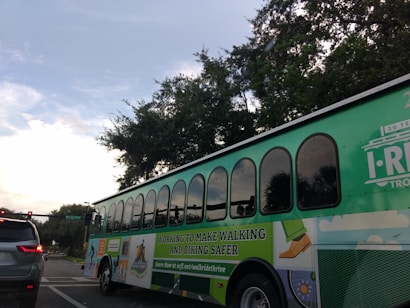 A large green trolley-style bus with multiple windows is seen on a city street. It bears signage promoting walking and biking safety, with a colorful design including images of people walking. A silver car is visible in the lane next to the bus, and there are traffic lights and trees in the background under a partly cloudy sky.