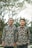 Two individuals stand side by side wearing matching patterned shirts with floral designs. The background features tall trees with a soft blur effect, suggesting an outdoor setting.