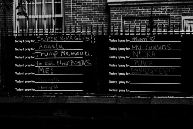 A blackboard with multiple lines starting with 'Today I pray for,' each followed by different handwritten messages in chalk expressing various prayers and wishes. The board is positioned against a brick wall with iron railings above it. The setting appears to be outdoors, possibly in an urban area.