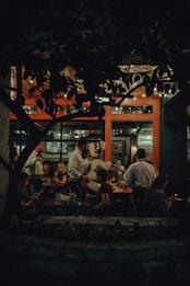 A cozy and dimly lit restaurant scene featuring a mariachi band performing for seated patrons. Customers are seated at wooden tables, enjoying their meals and drinks, while the musicians, dressed in traditional attire, entertain with string instruments. The interior has warm lighting and natural elements, with a prominent plant casting shadows against a backdrop of large windows.