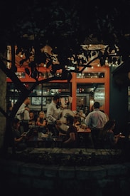 A cozy and dimly lit restaurant scene featuring a mariachi band performing for seated patrons. Customers are seated at wooden tables, enjoying their meals and drinks, while the musicians, dressed in traditional attire, entertain with string instruments. The interior has warm lighting and natural elements, with a prominent plant casting shadows against a backdrop of large windows.