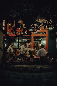 A cozy and dimly lit restaurant scene featuring a mariachi band performing for seated patrons. Customers are seated at wooden tables, enjoying their meals and drinks, while the musicians, dressed in traditional attire, entertain with string instruments. The interior has warm lighting and natural elements, with a prominent plant casting shadows against a backdrop of large windows.