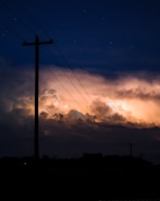 An electric pole silhouetted against a dramatic sky filled with glowing clouds and flashes of lightning. The scene is captured during nighttime with a palette of deep blues and bright, fiery oranges lighting up the horizon.