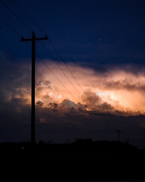 An electric pole silhouetted against a dramatic sky filled with glowing clouds and flashes of lightning. The scene is captured during nighttime with a palette of deep blues and bright, fiery oranges lighting up the horizon.