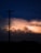 An electric pole silhouetted against a dramatic sky filled with glowing clouds and flashes of lightning. The scene is captured during nighttime with a palette of deep blues and bright, fiery oranges lighting up the horizon.