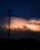 An electric pole silhouetted against a dramatic sky filled with glowing clouds and flashes of lightning. The scene is captured during nighttime with a palette of deep blues and bright, fiery oranges lighting up the horizon.