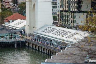 A riverside dining area lined with people, situated next to a large building with signs reading 'Felon's Brewing Co'. The setting includes modern architecture with geometric designs, surrounded by water. Trees and city streets with traffic are visible in the background.