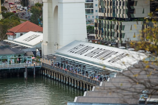 A riverside dining area lined with people, situated next to a large building with signs reading 'Felon's Brewing Co'. The setting includes modern architecture with geometric designs, surrounded by water. Trees and city streets with traffic are visible in the background.
