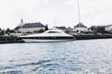 A sleek, modern yacht is moored along a waterfront with a historic building featuring a green copper dome in the background. The scene is set against a lightly clouded sky and the water appears calm with small ripples.