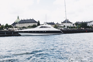 A sleek, modern yacht is moored along a waterfront with a historic building featuring a green copper dome in the background. The scene is set against a lightly clouded sky and the water appears calm with small ripples.