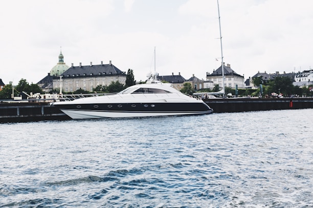 A sleek, modern yacht is moored along a waterfront with a historic building featuring a green copper dome in the background. The scene is set against a lightly clouded sky and the water appears calm with small ripples.