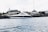 A sleek, modern yacht is moored along a waterfront with a historic building featuring a green copper dome in the background. The scene is set against a lightly clouded sky and the water appears calm with small ripples.
